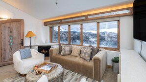 A cozy living room with a fireplace, a sofa, a coffee table, and a view of a snowy landscape through large windows. - Penthouse At The Peaks 826 (Mountain Village)