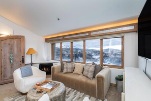 A cozy living room with a fireplace, a sofa, a coffee table, and a view of a snowy landscape through large windows.
