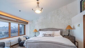 A bedroom with a large bed, two bedside lamps, a view of mountains through a window, and a small round table. - Penthouse At The Peaks 826 (Mountain Village)
