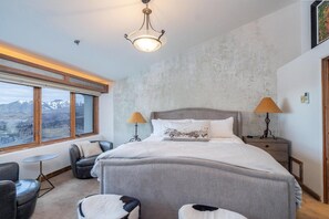 A bedroom with a large bed, two bedside lamps, a view of mountains through a window, and a small round table.