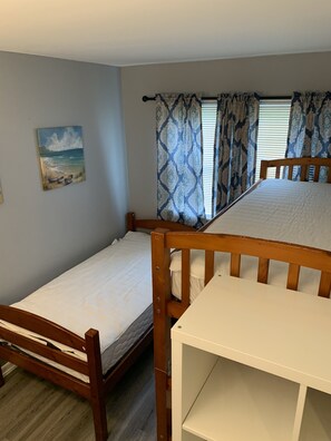 Cozy sleeping quarters with a single bed and bunk bed arrangement, featuring a window with patterned curtains and a beach-themed painting.