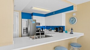 A modern kitchen with white cabinets, a stainless steel refrigerator, a microwave, and a coffee maker. There are two blue barstools and a clock on the wall. - IP 208: Sunglasses Required - Your Beachfront Sanctuary Starts Here! (Fort Walton Beach)