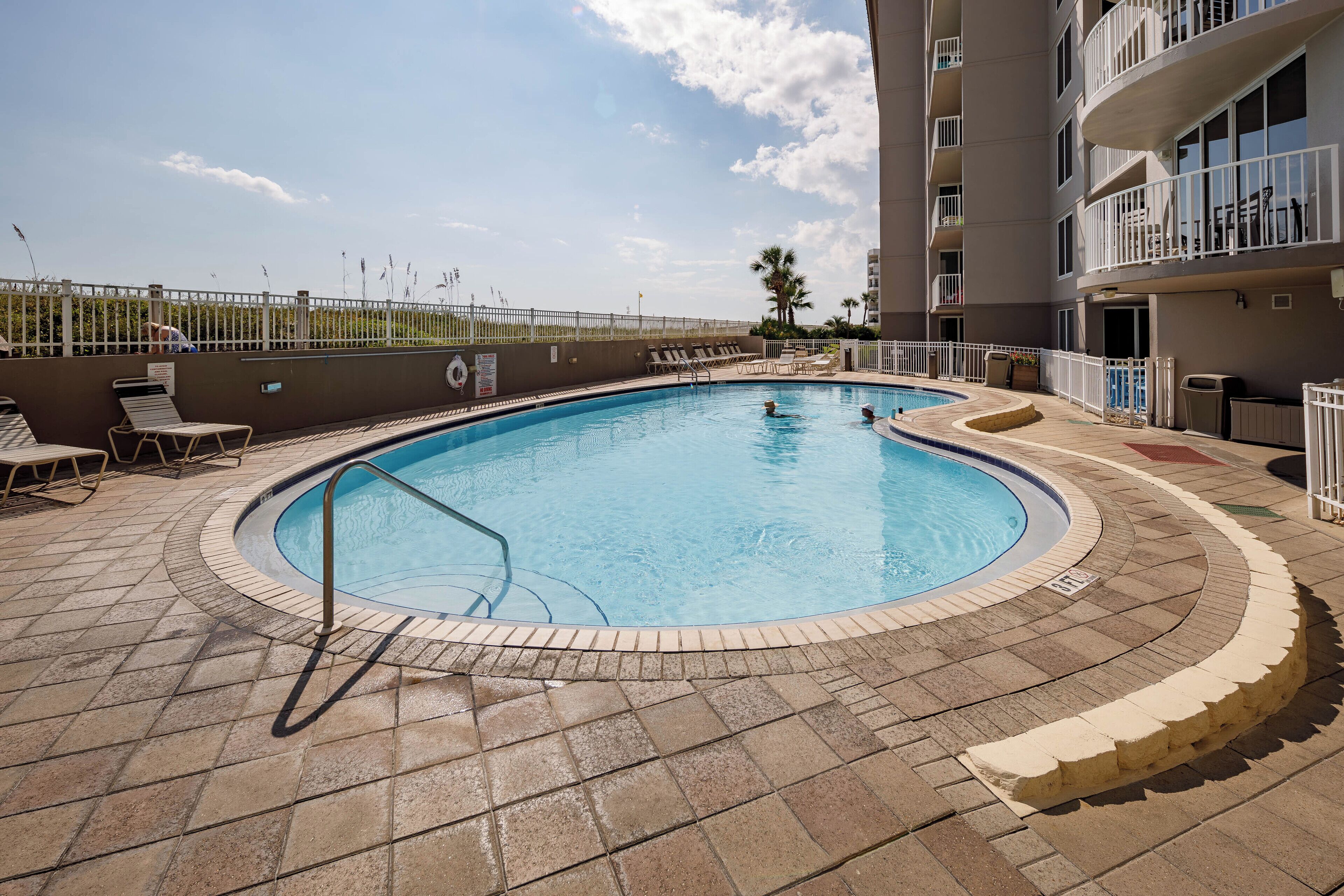 A swimming pool with a tiled deck, surrounded by lounge chairs and a fence.