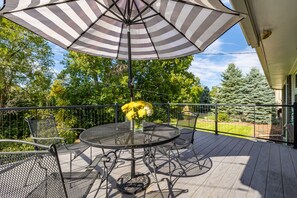 Eine Terrasse mit einem runden Tisch und Stühlen, einem gestreiften Sonnenschirm und einem Blumenstrauß mit gelben Blumen.
