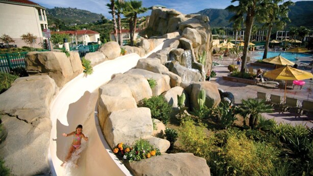 Un tobogán de agua con rocas grandes, una zona de piscina con sombrillas y palmeras.