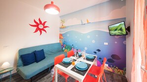 Ein Zimmer mit einem blauen Sofa, einem Esstisch mit orangefarbenen Stühlen, einer roten Blütenwanddekoration und einem an der Wand befestigten Fernseher.