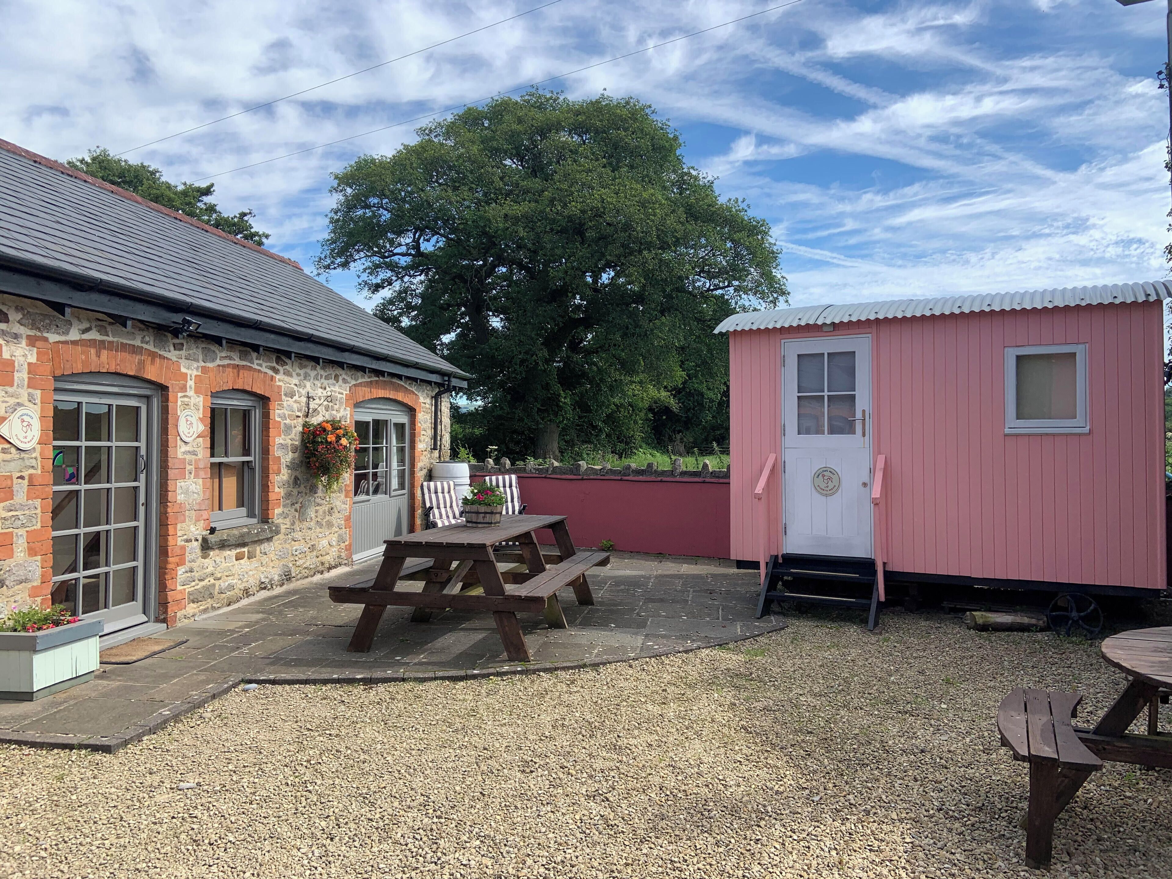 A stone building with a red door and a pink shed. There are picnic tables and a gravel area.