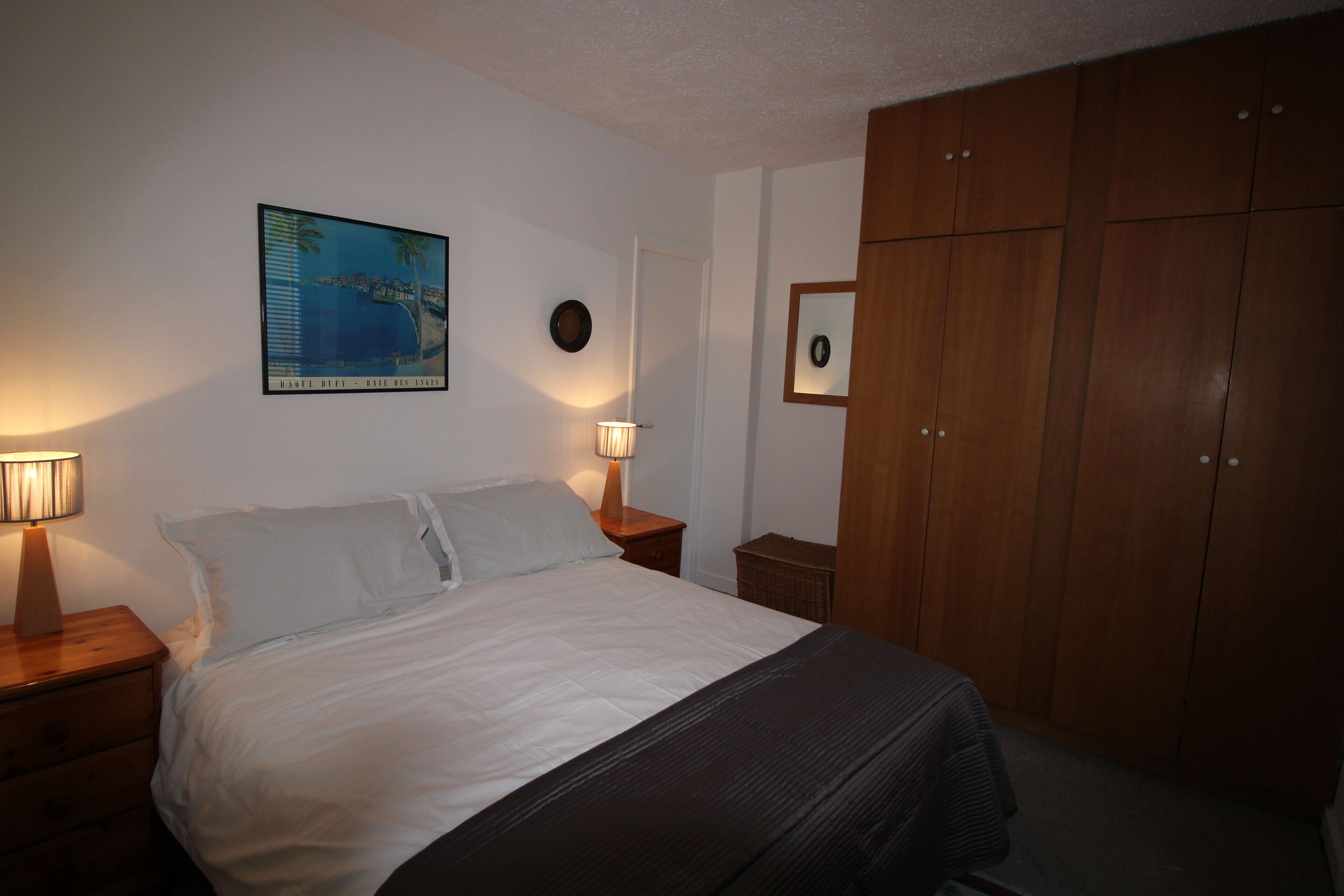 Un dormitorio con cama, dos lámparas de noche, un armario de madera, un cuadro en la pared y otro cuadro sobre el armario.