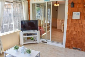 A room with a TV on a white stand, a small table with plants, and a sliding glass door leading to a balcony.