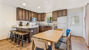 Una cocina con armarios de madera, microondas, refrigerador, mesa de comedor con sillas y un cubo de basura.