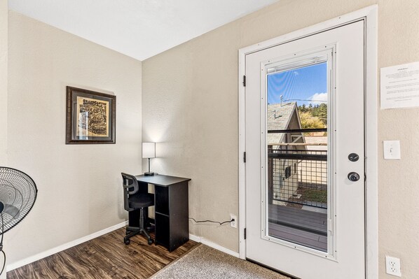 A room with a door leading to a balcony, a desk with a chair, a fan, and a framed picture on the wall.