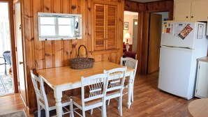 A wooden-paneled kitchen with a wooden table and white chairs, a refrigerator, and a window with blinds. - Charming Ranch Home with Sunroom & Fenced Yard – Perfect Cape Escape! (Dennis Port)