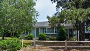 A single-story house with a front porch, surrounded by mature trees and a wooden fence. - Charming Ranch Home with Sunroom & Fenced Yard – Perfect Cape Escape! (Dennis Port)
