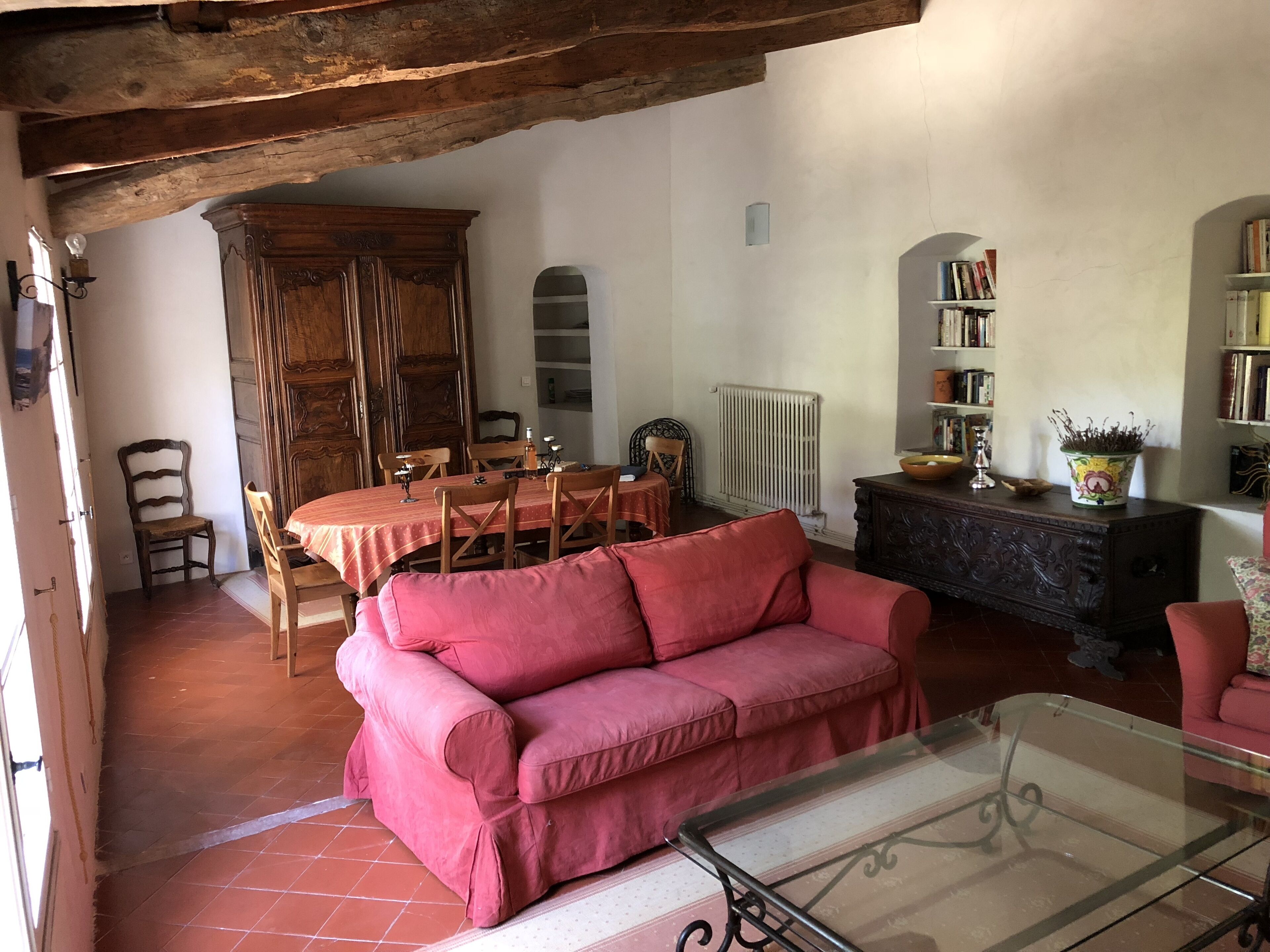 Un salón acogedor con un sofá rojo, una mesa de centro de cristal y una mesa de comedor de madera con sillas. Se observa una estantería y un radiador al fondo.