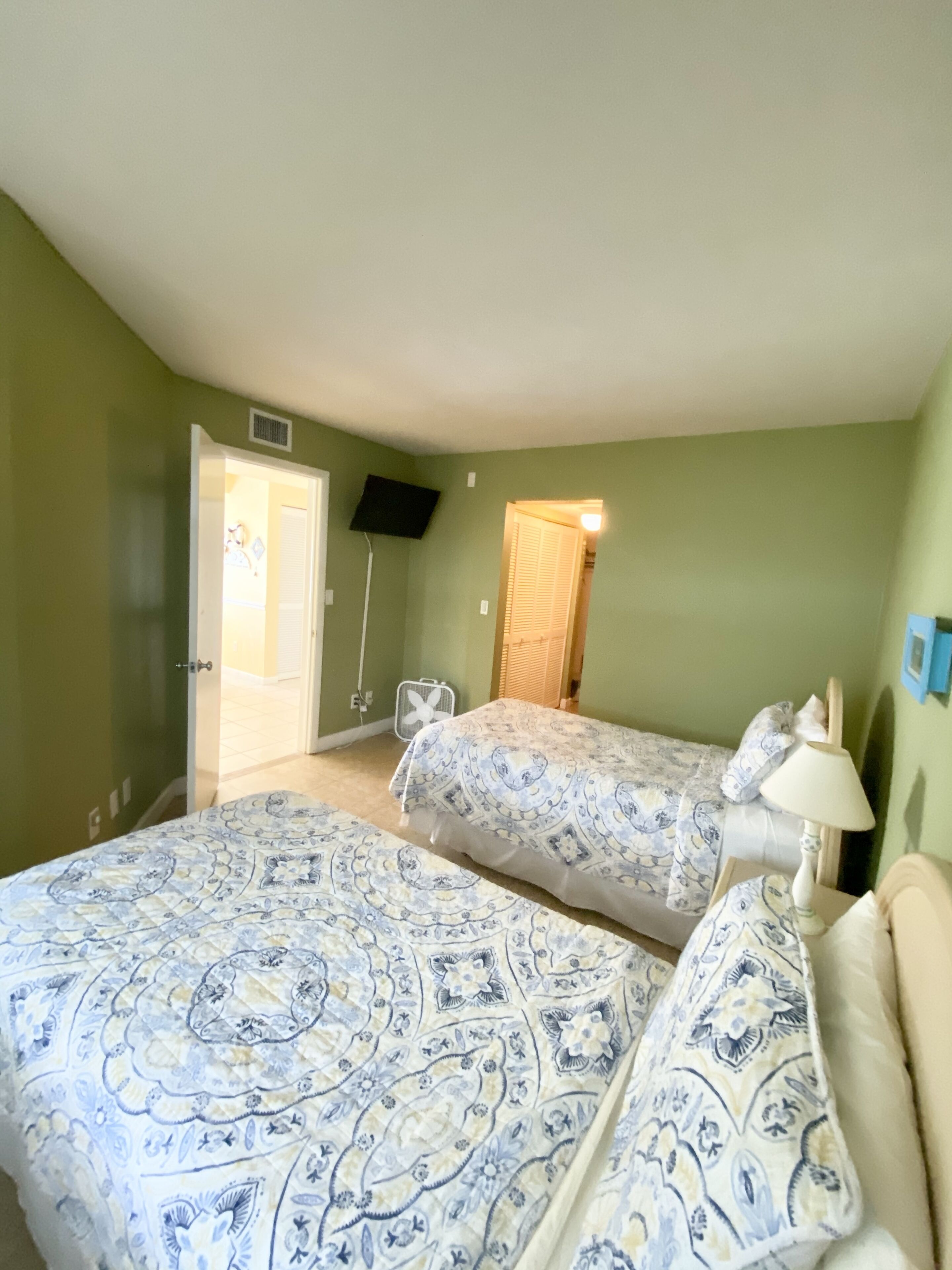 Ein Zimmer mit zwei Betten, einem an der Wand befestigten Fernseher, einer Tür und einem Fenster mit Jalousien.