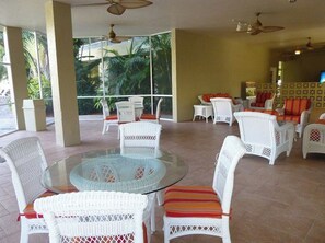 A spacious outdoor seating area with white wicker furniture and glass tables, surrounded by large windows offering views of greenery.