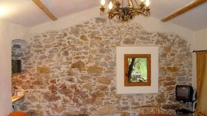 A stone-walled room with a dining table, chairs, a television, and a window with a view of greenery.