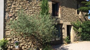 Un bâtiment en pierre à l’aspect rustique, avec une porte en bois et un petit jardin agrémenté de plantes en pot et d’un banc.