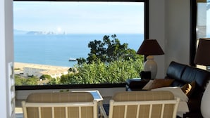 A room with a large window offering a view of the sea and distant buildings. There is a black leather sofa and two chairs with a small table and lamp between them. - Villa in Begur with sea views-Capacity 9 people.-A/C.Pool. BEGUR-COSTA BRAVA (BEGUR)
