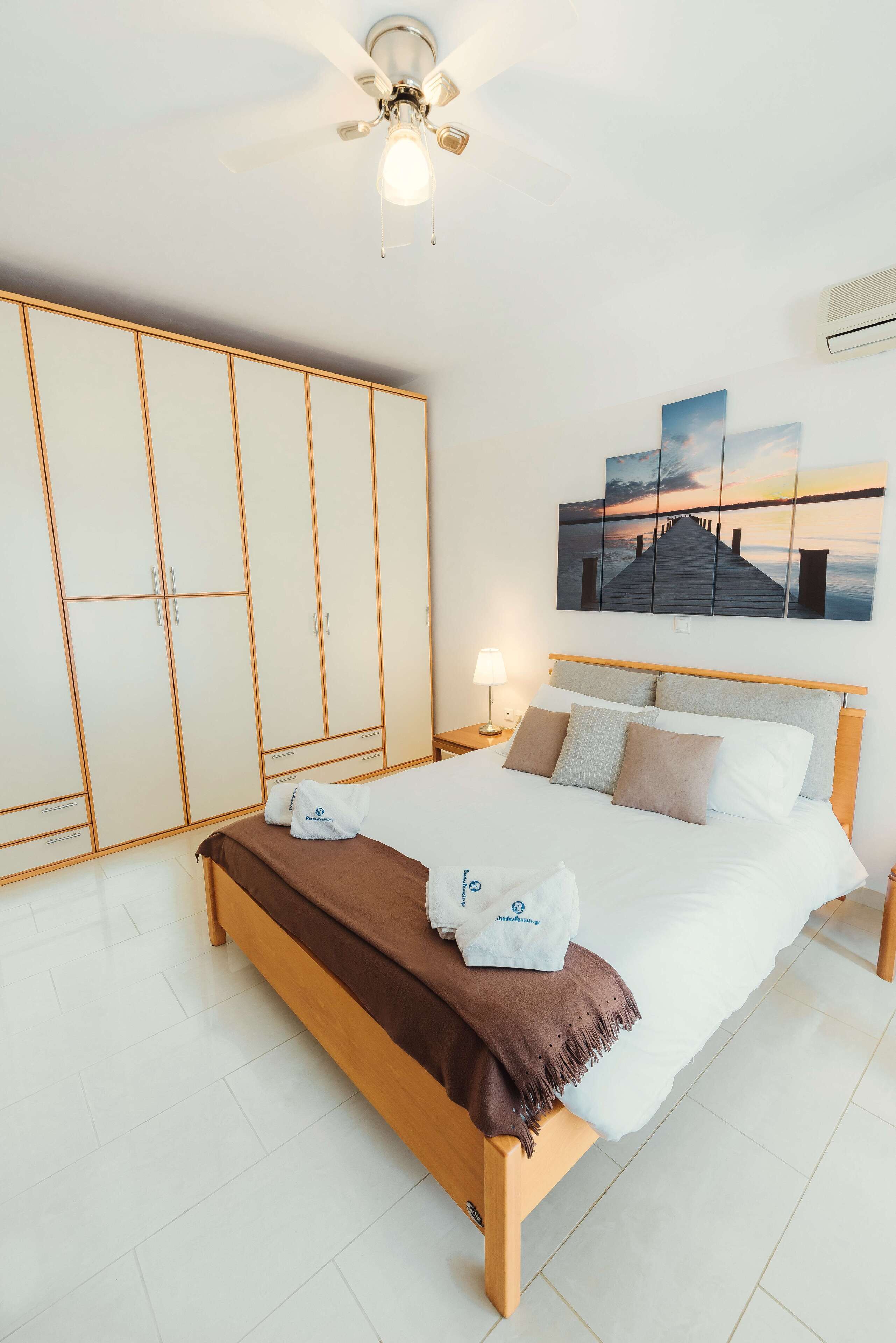 A bedroom with a bed, a wardrobe, a ceiling fan, a nightstand, a lamp, and a painting on the wall.