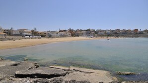 Una zona costera con playa de arena y agua cristalina. Hay edificios a lo largo de la orilla, y algunas personas se ven a lo lejos.