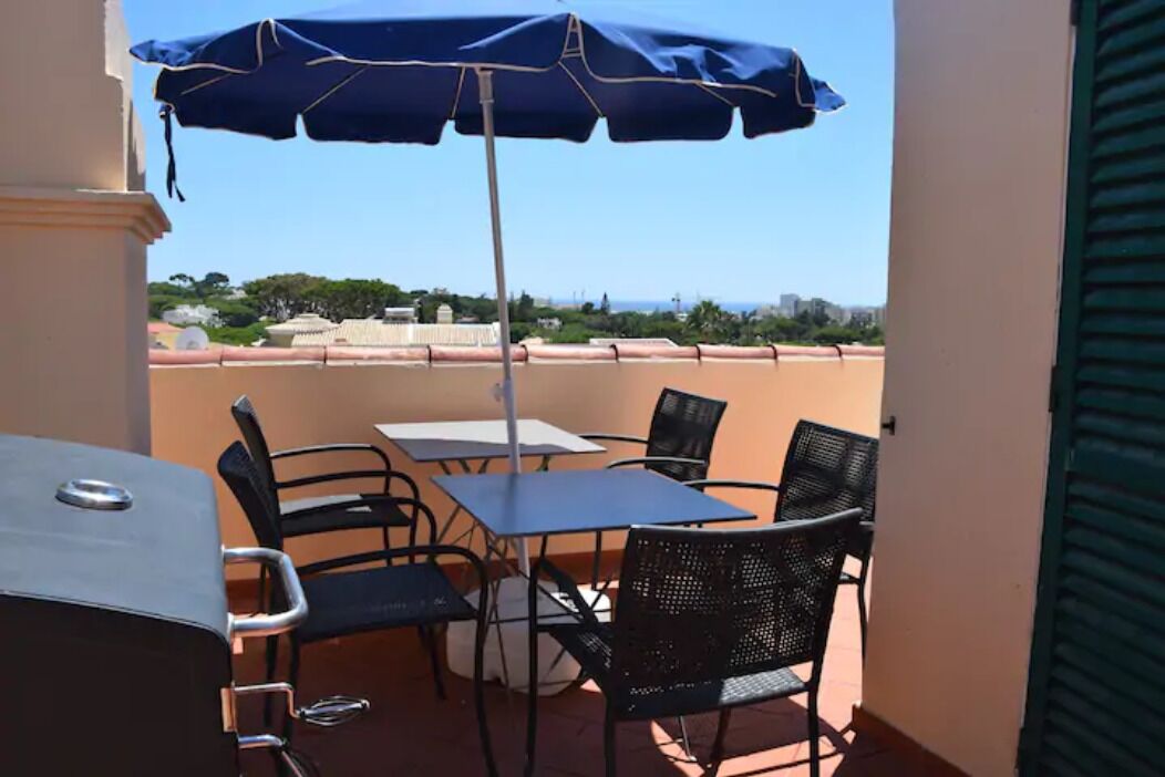 A balcony with a table and chairs, a grill, and a blue umbrella.