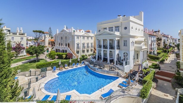Um hotel com piscina rodeada por espreguiçadeiras e guarda-sóis, um jardim bem cuidado e um céu azul límpido.