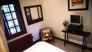Un dormitorio con cama, una silla, un televisor sobre un mueble, un radiador y una ventana con vistas a la vegetación.