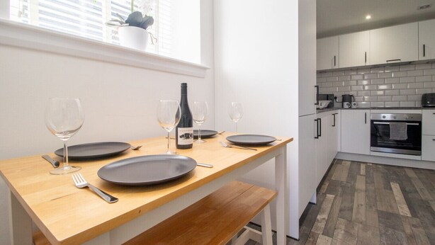 A modern kitchen with white cabinets, a wooden dining table set for four, and a window with a potted plant. - Abbey House Apartment A (Paisley)
