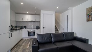 A modern kitchen with white cabinets, a built-in oven, and a microwave. There is a black leather sofa in the living area, a remote control on the sofa, and a staircase leading to another level. - Abbey House Apartment A (Paisley)