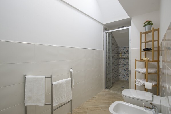 A modern bathroom with a shower, toilet, and sink. There is a towel rack with towels, a wooden shelf with toiletries, and a potted plant.