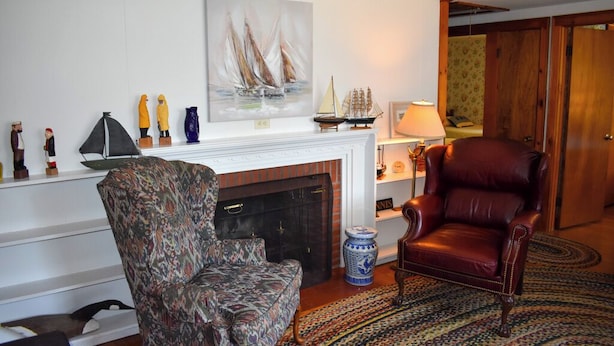 A living room with a patterned armchair, a leather armchair, a fireplace, and a painting of sailboats. - Comfortable, Cape Cod home is the perfect place to unwind this summer (West Dennis)