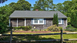 A single-story house with a grey shingle roof, white window shutters, and a small front porch. - Comfortable, Cape Cod home is the perfect place to unwind this summer (West Dennis)