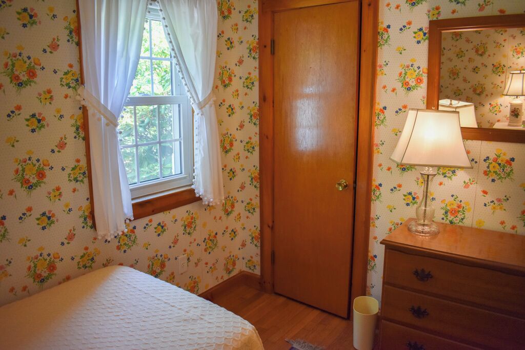 A bedroom with floral wallpaper, a wooden door, a bed with a white bedspread, a dresser with a lamp, and a window with white curtains.