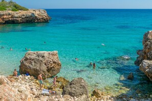 A rocky coastline with clear turquoise water, people swimming and sunbathing, and a cliff with vegetation. - CALA RAFALINO - Chalet with terrace in the peaceful area of Cala Morlanda. Free WiFi (Manacor)