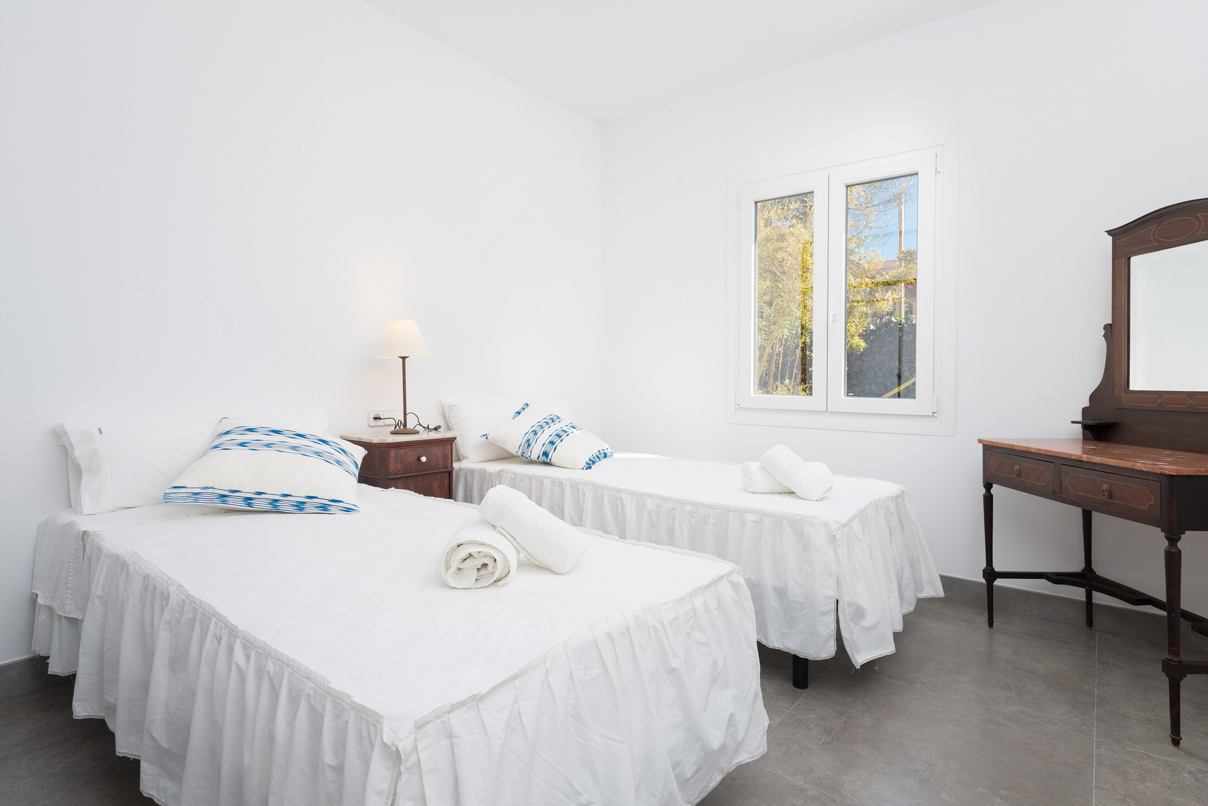 A hotel room with two beds, a desk, a nightstand, and a window with a view of trees.