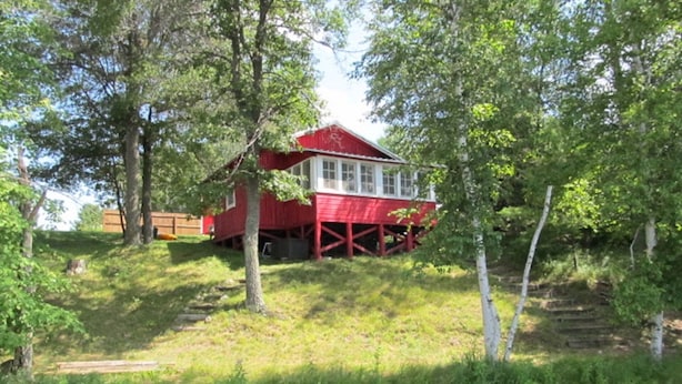A red house with a white window, surrounded by trees and a small stream.