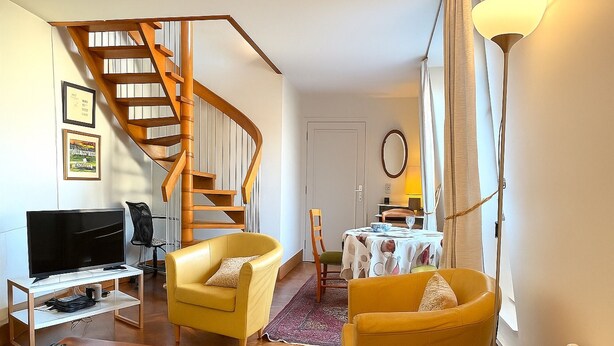 A living room with a wooden staircase, yellow armchairs, a television, and a dining area with a table and chairs. - Le Penthouse Thorigny (Paris)