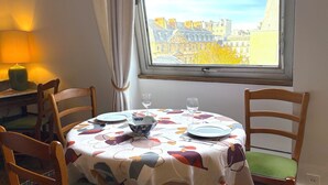 A dining table with a floral tablecloth, two chairs, a window with a view of buildings, and a lamp on a side table. - Le Penthouse Thorigny (Paris)