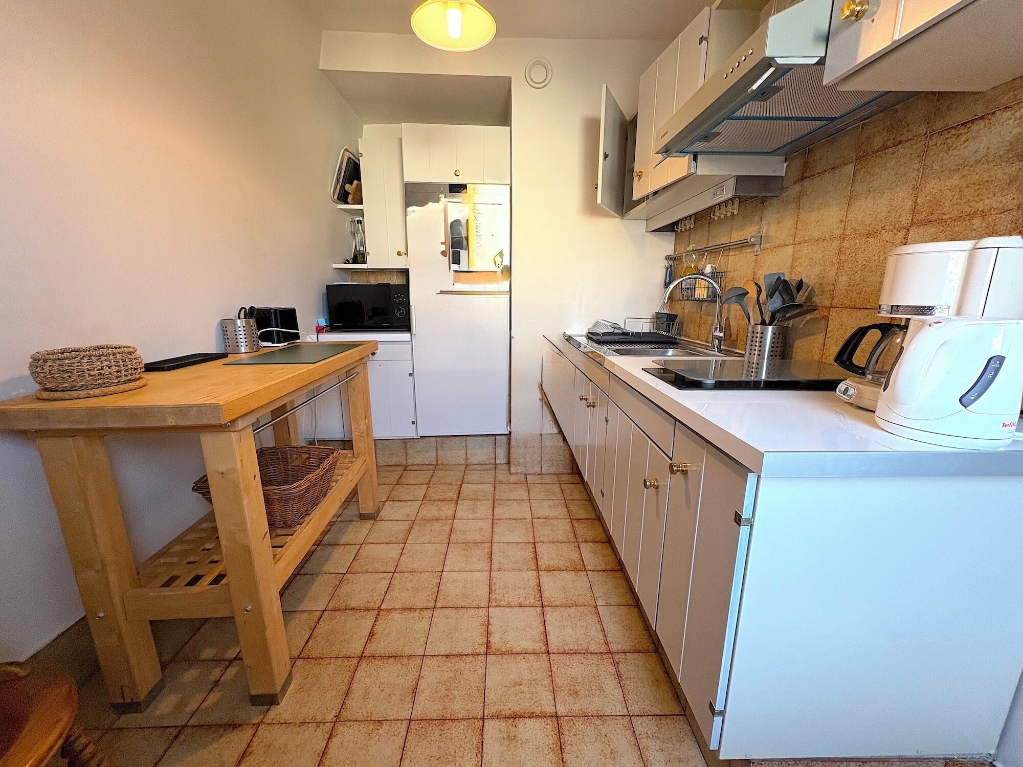 Una cucina compatta con mobili bianchi, un tavolo di legno, un forno a microonde, una macchina da caffè e un pavimento piastrellato.