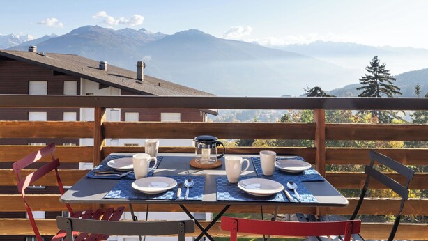 A table set for two with a mountain view, wooden deck, and red chairs.