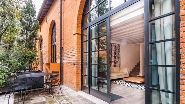 Une maison moderne avec une façade en briques, de grandes portes vitrées donnant sur un espace de vie comprenant un canapé et un escalier.