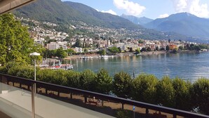 A panoramic view of a lakeside city with mountains in the background, a marina with boats, and a pedestrian walkway along the water.