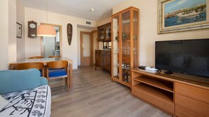 Un salon avec un téléviseur, des meubles en bois, un coin repas avec des chaises et, au fond, une cuisine.