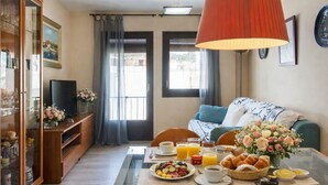 Une pièce bien éclairée, avec une table à manger dressée pour le petit-déjeuner, un canapé agrémenté d’un arrangement floral, un téléviseur posé sur un meuble et une grande fenêtre avec des rideaux.