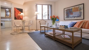 En moderne stue med sofa, sofabord og stole. Der er et vindue med persienner, et billede på væggen og et køkkenområde i baggrunden.