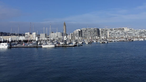 Un port de plaisance où sont amarrés de nombreux bateaux, une tour imposante et, au loin, des bâtiments modernes.
