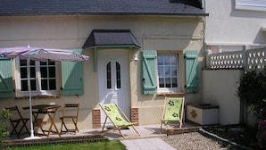 Une maison avec un porche à l’avant, une porte et deux fenêtres avec des volets. On trouve deux chaises de jardin et une table sur le porche.