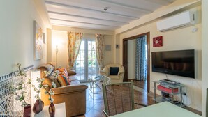 A living room with a sofa, a TV, a table, and a chair. There is a window with curtains and a door leading to another room. - La Quinta de Isla Canela II - Isla Canela Golf GQHII2P2 (Isla Canela)