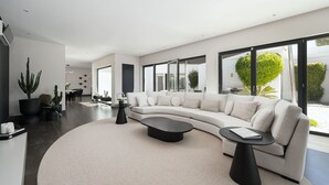 A modern living room with a large L-shaped sofa, a round coffee table, and a small side table. There are potted plants and a dining area visible in the background. - Villa Allegra - Quinta do Lago (Quinta do Lago)